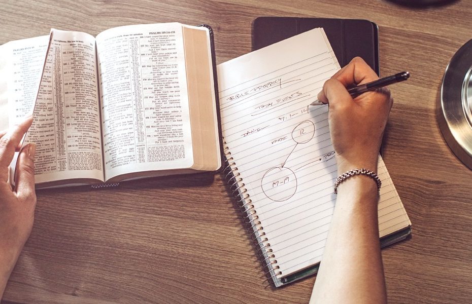 Mãos escrevendo anotações em um caderno ao lado da Bíblia aberta